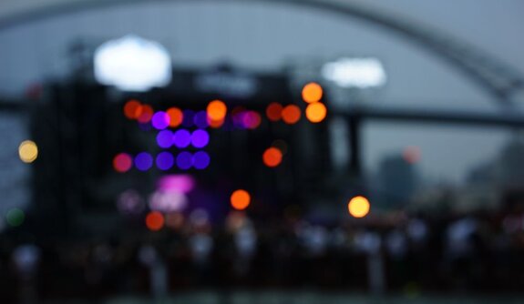 Foto desfocada (bokeh) de um palco de show ao entardecer, com luzes coloridas em roxo, laranja e amarelo, estrutura de arco metálico ao fundo e silhuetas do público em primeiro plano.
