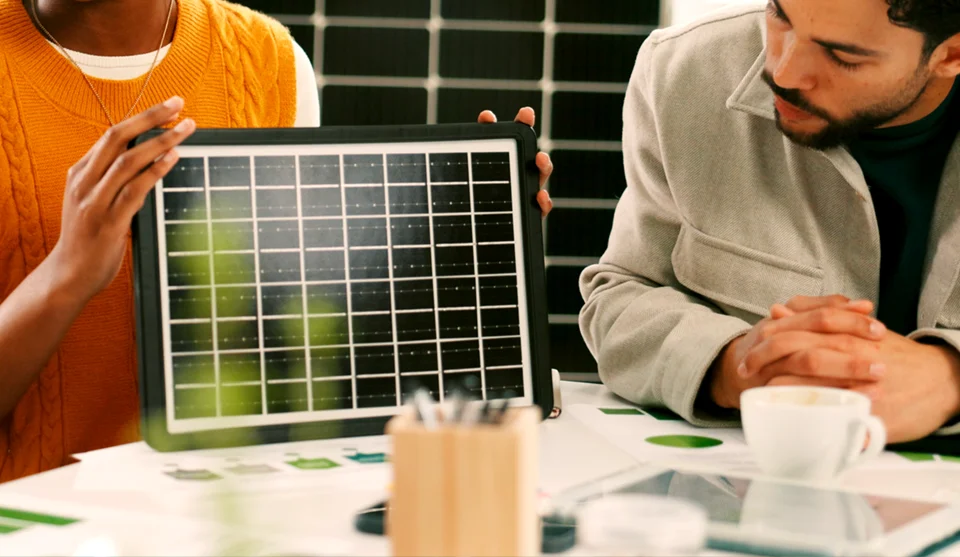 Mulher segurando um protótipo de placa solar em miniatura em cima de uma mesa de escritório com vários objetos em cima da mesa. Homem adulto ao lado direito da imagem olhando a placa solar.