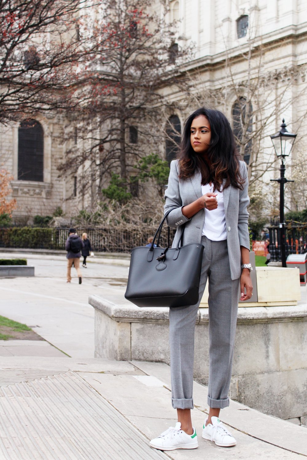 Sachini wearing a grey suit and white Adidas sneakers holding a black bag