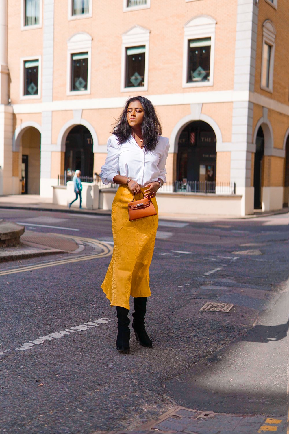 Sachini standing in the street with a white blouse, long yellow skirt, black boot holding a brown Hermès mini Kelly