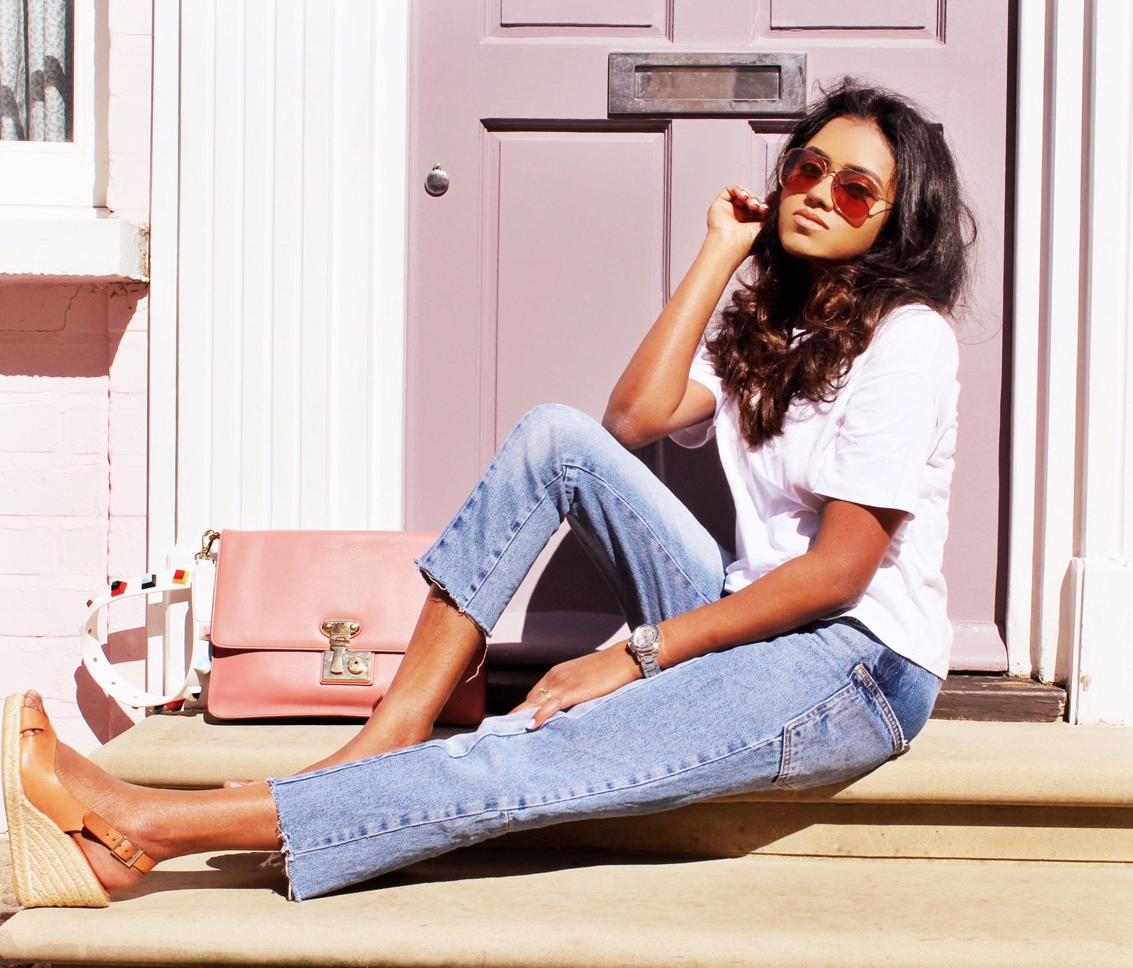 Sachini sitting in front of a pink door wearing a white t-shirt, blue jeans with a pink Dolce and Gabanna