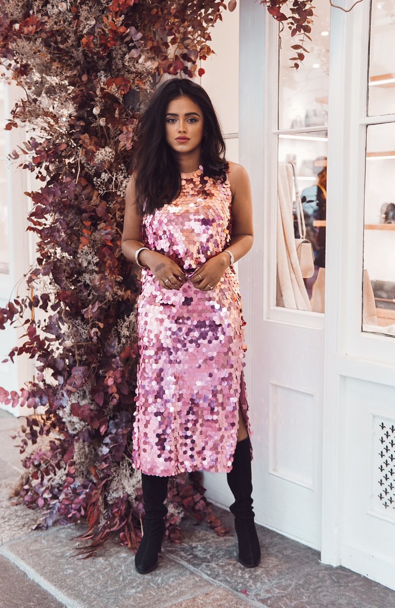 Sachini wearing a pink Chi Chi London sequin dress and high black boots standing in front of a shop next to a tree with red leaves