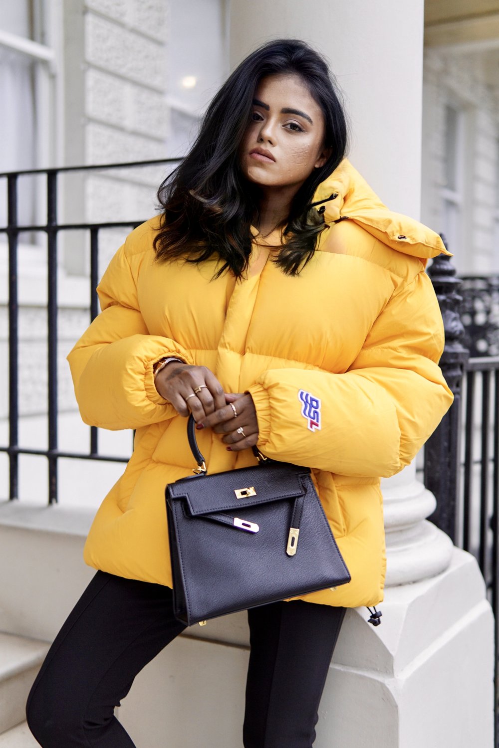 Sachini wearing a yellow Diesel puffer jacket holding a black Hermès Kelly bag