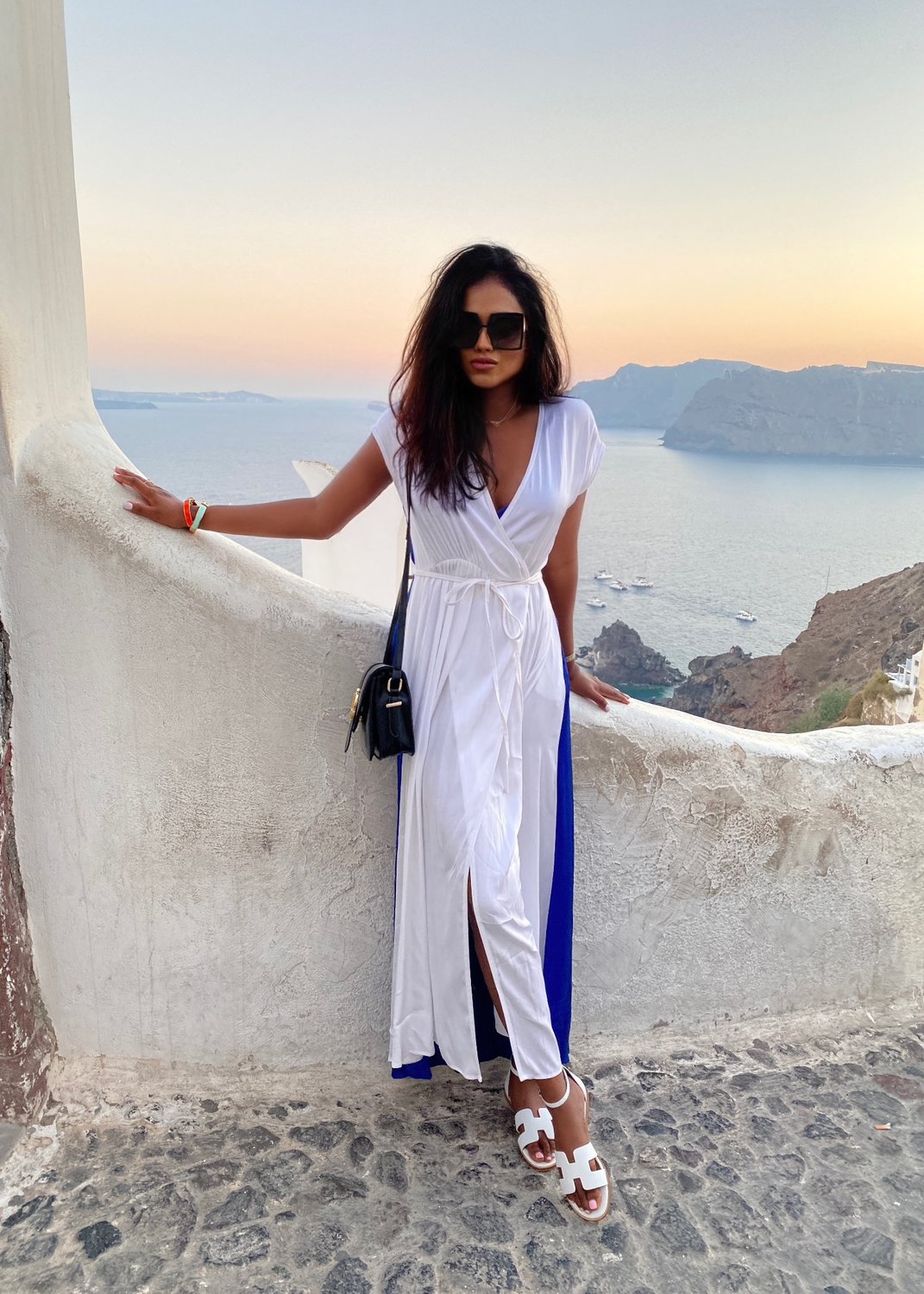 Sachini wearing sunglasses, a blue and white Ioanna Kourbela dress and white Hermes sandals with in the background a sea and sunset