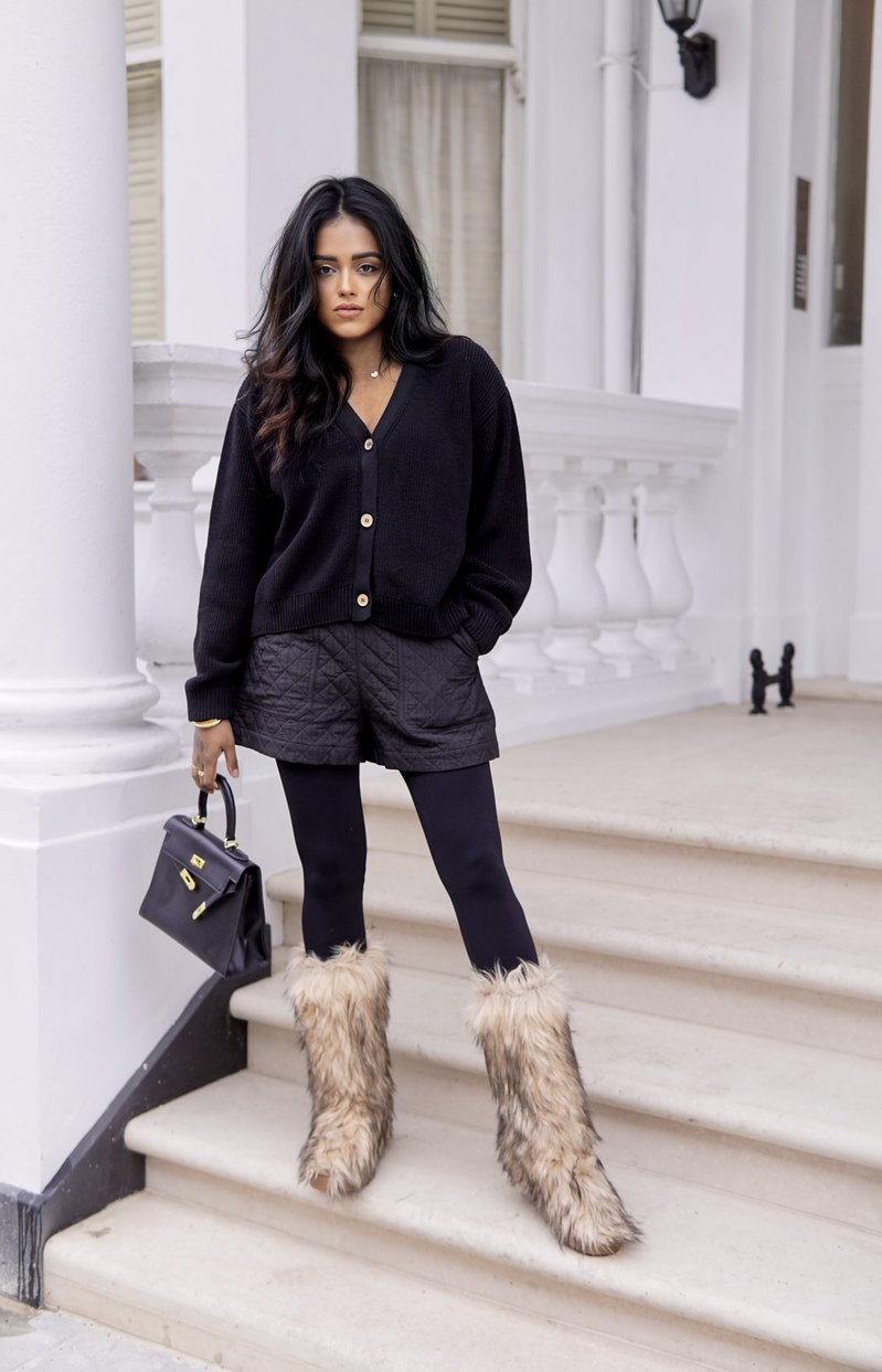 Sachini wearing a black top, shorts and leggings with faux fur boots holding a black Hermès Kelly bag