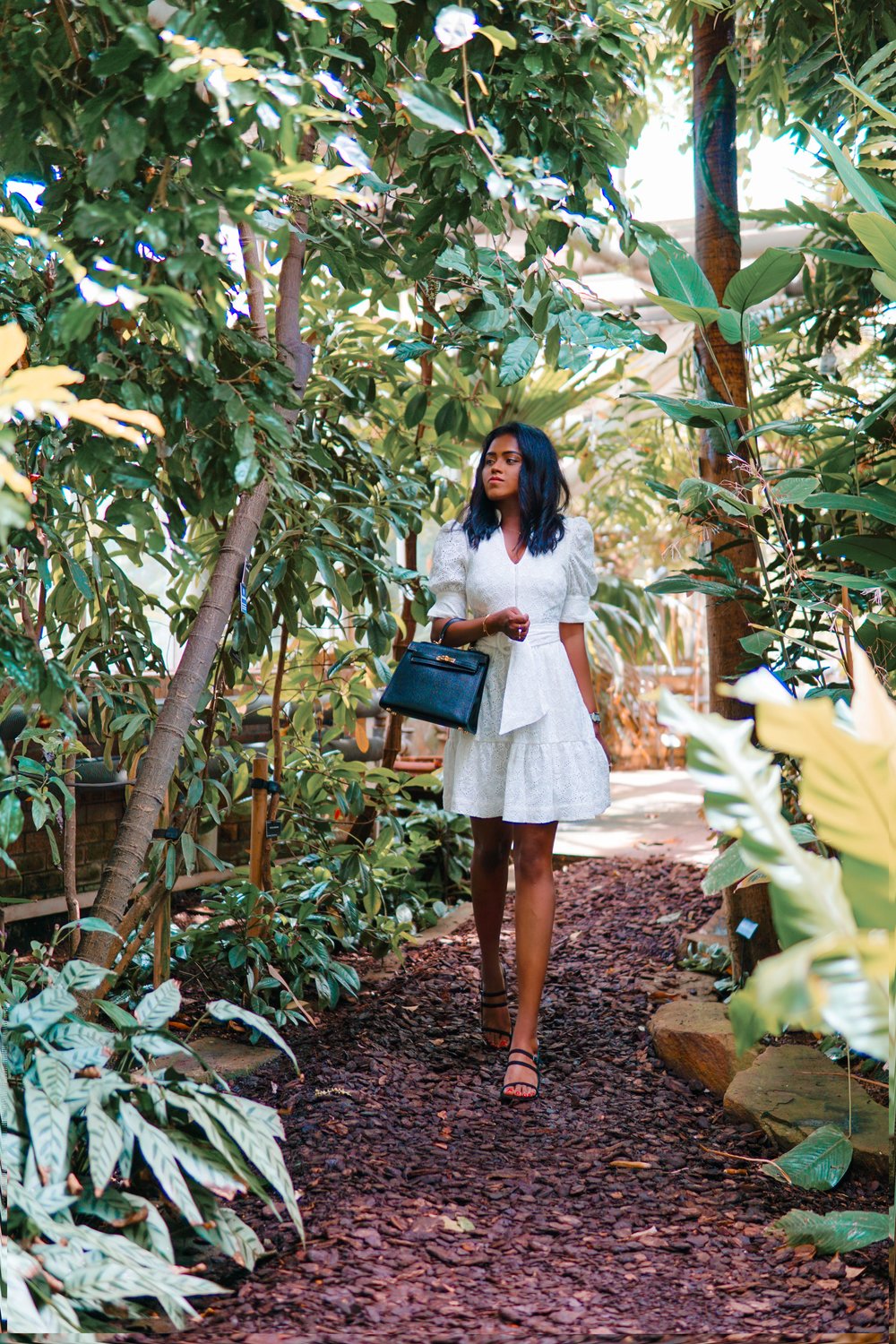 Sachini wearing a white dress holding a black Hermès Kelly bag walking in a botanic garden