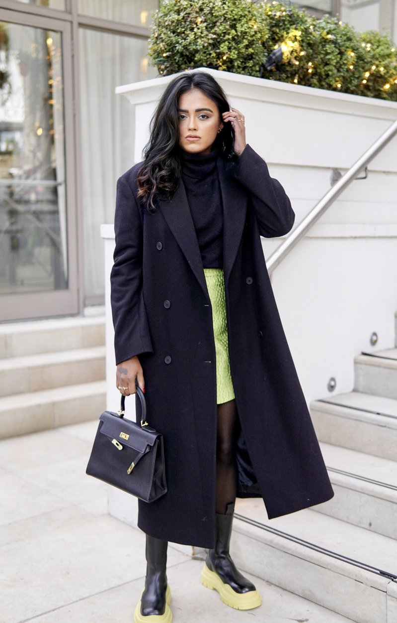 Sachini standing in front of a stair wearing a black coat, a light green skirt, a black Hermès Kelly bag and black and green Bottega Veneta boots