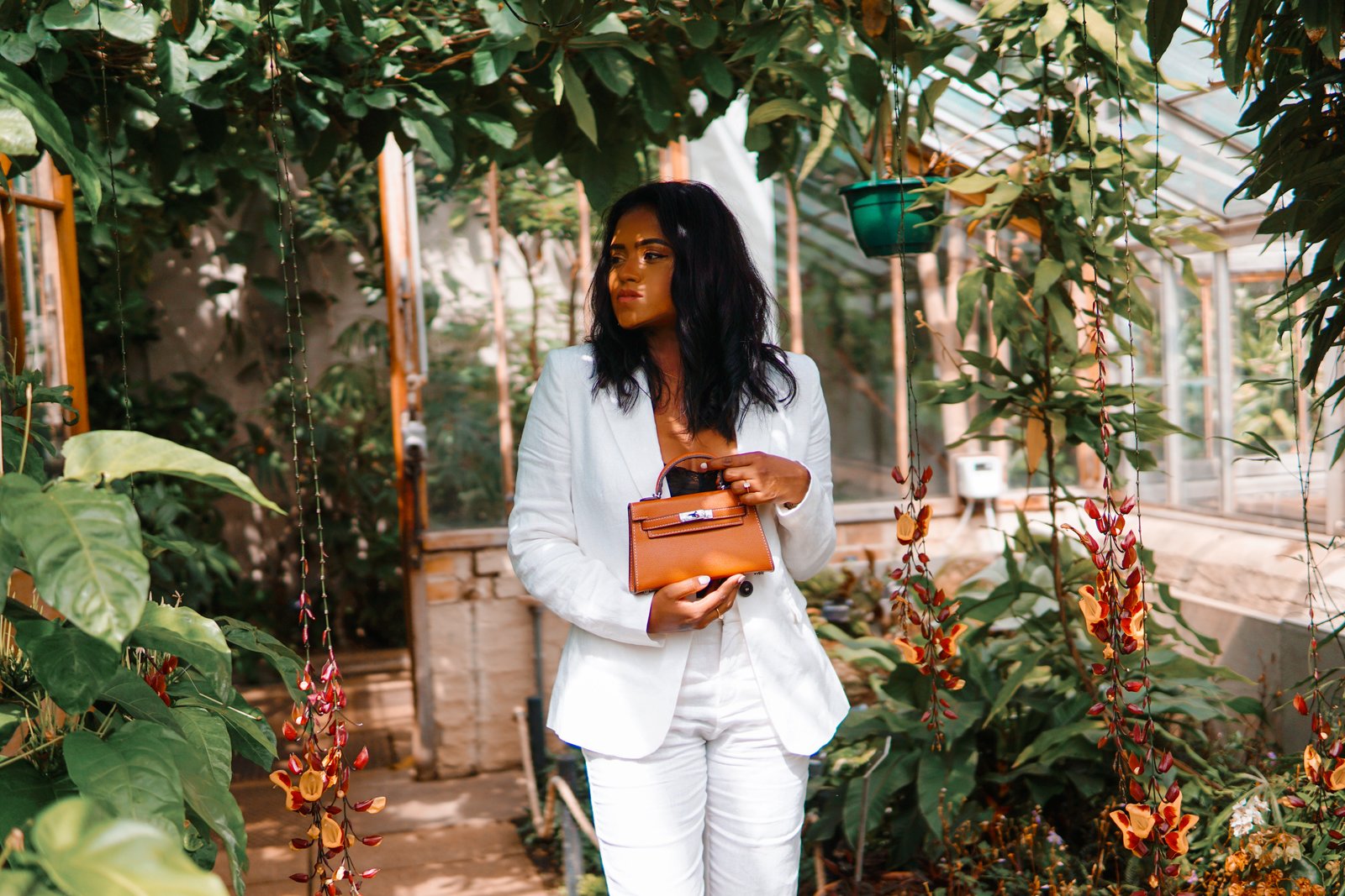 Sachini standing between exotic trees wearing a white suit holding a brown Hermès mini Kelly bag