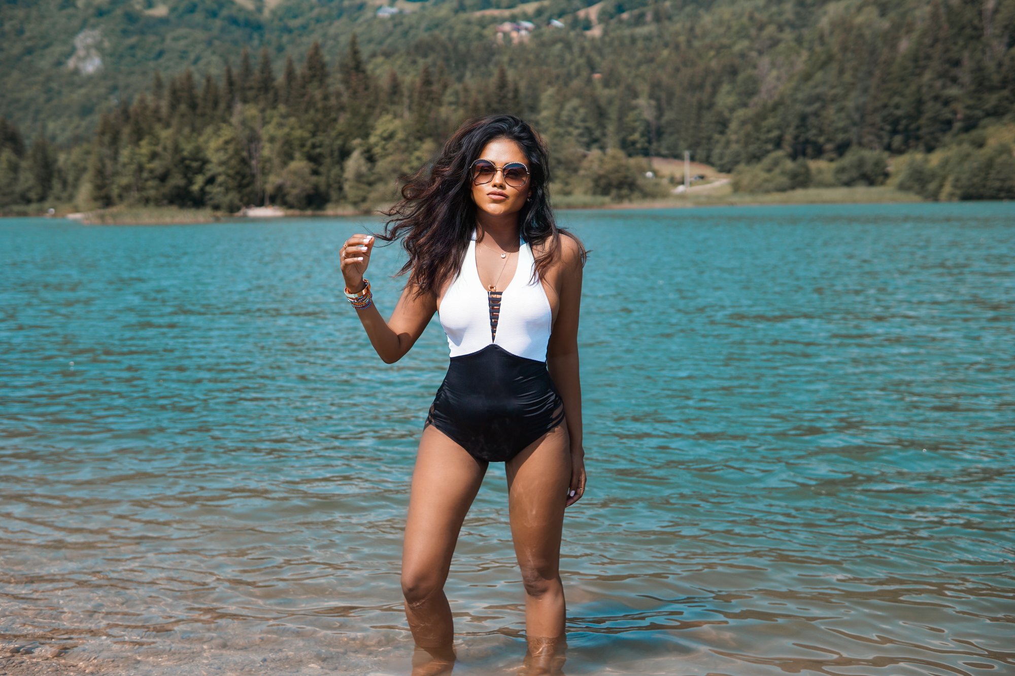 Sachini wearing Chanel sunglasses and a black and white swimsuit standing in front a mountain lake