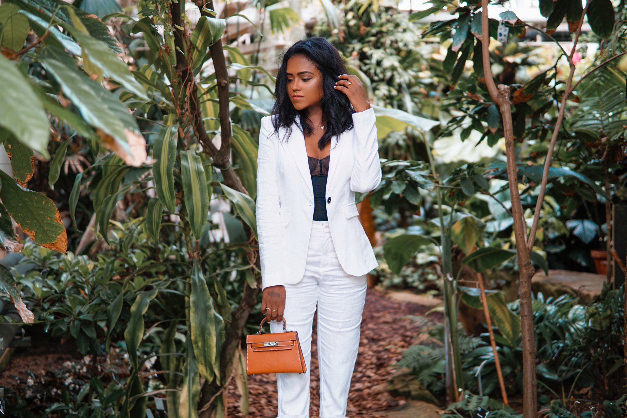 Sachini standing between exotic trees wearing a white suit holding a brown Hermès mini Kelly bag