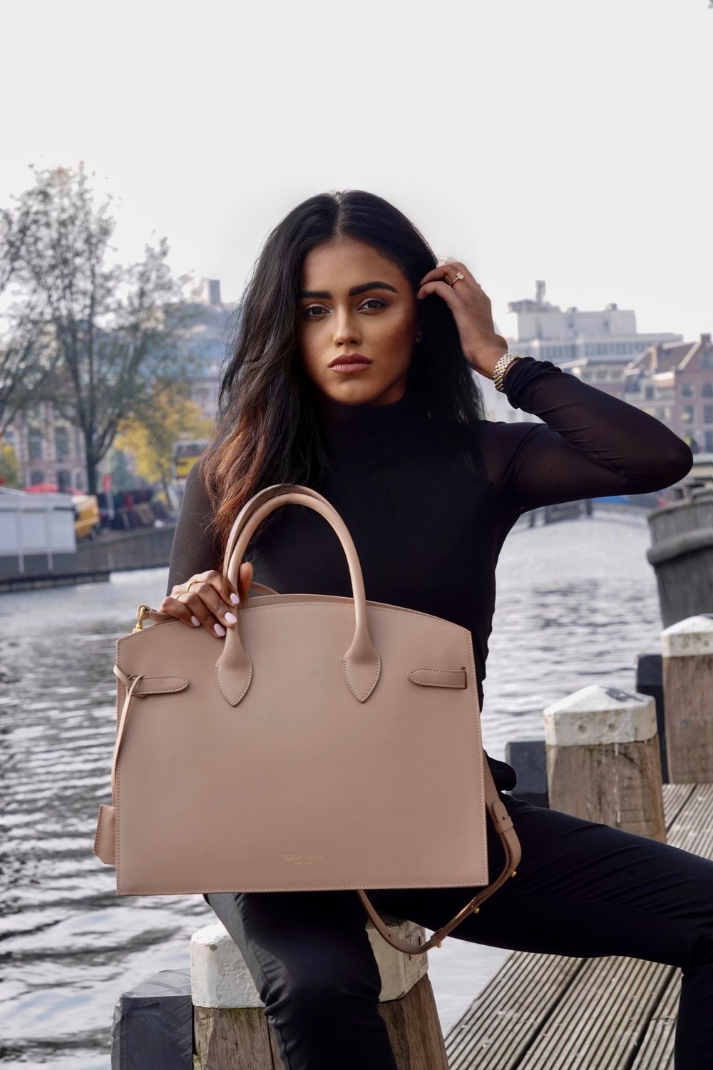 Close up of sachini sitting next to a canal in Amsterdam wearing a black top and holding a beige Teddy Blake bag