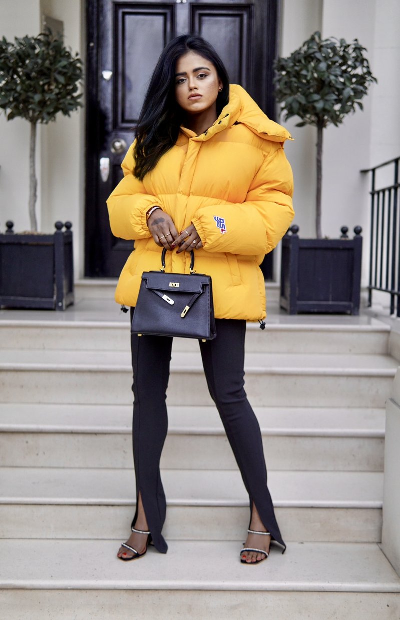 Sachini standing in front of a stair wearing a yellow Diesel puffer jacket and black Amina Muaddi shoes holding a black Hermès Kelly bag
