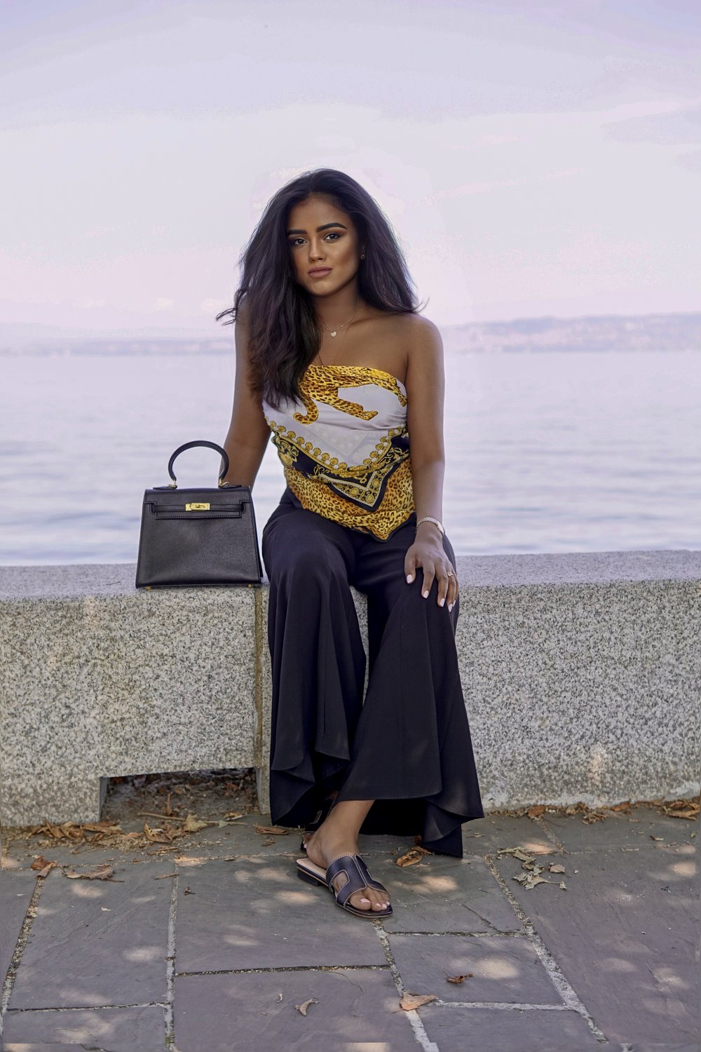 Sachini sitting in front of the lake of Geneva with black trousers and an Hermès scarf as a top with a black Hermès Kelly next to her