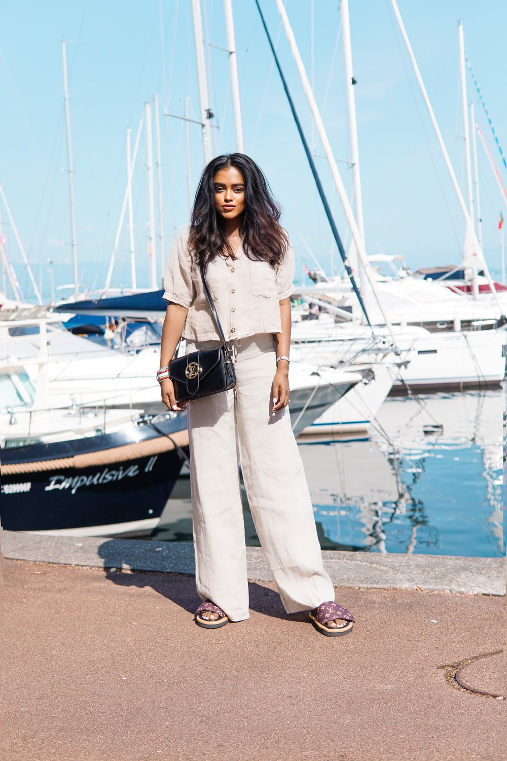 Sachini at a harbour in front of boots wearing a beige top and trousers and Louis Vuitton slides holding a black Louis Vuitton bag