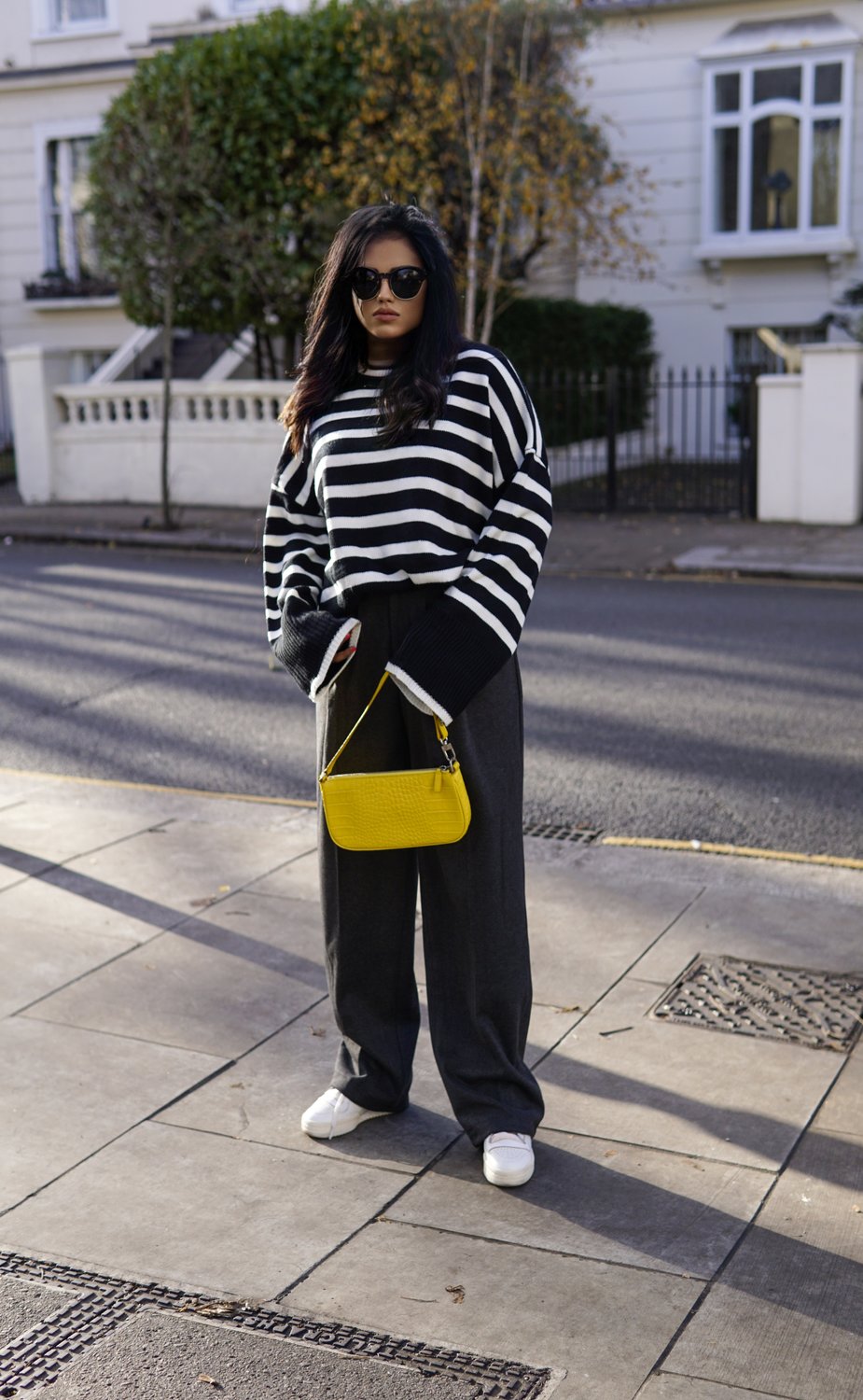 Sachini standing on a sidewalk wearing an oversized Chicwish striped jumper and trousers with a yellow bag