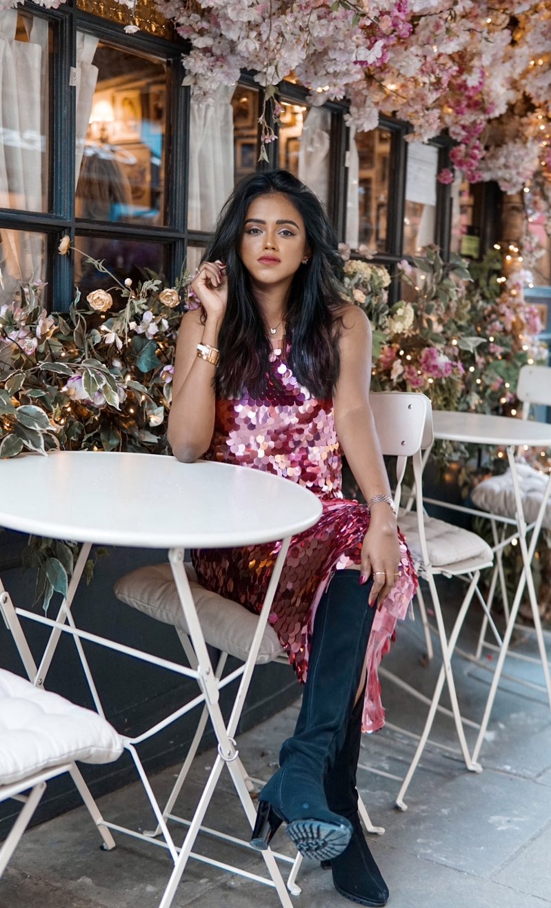 Sachini sitting down at a cafe wearing a Chi Chi London pink sequin dress and high black boots