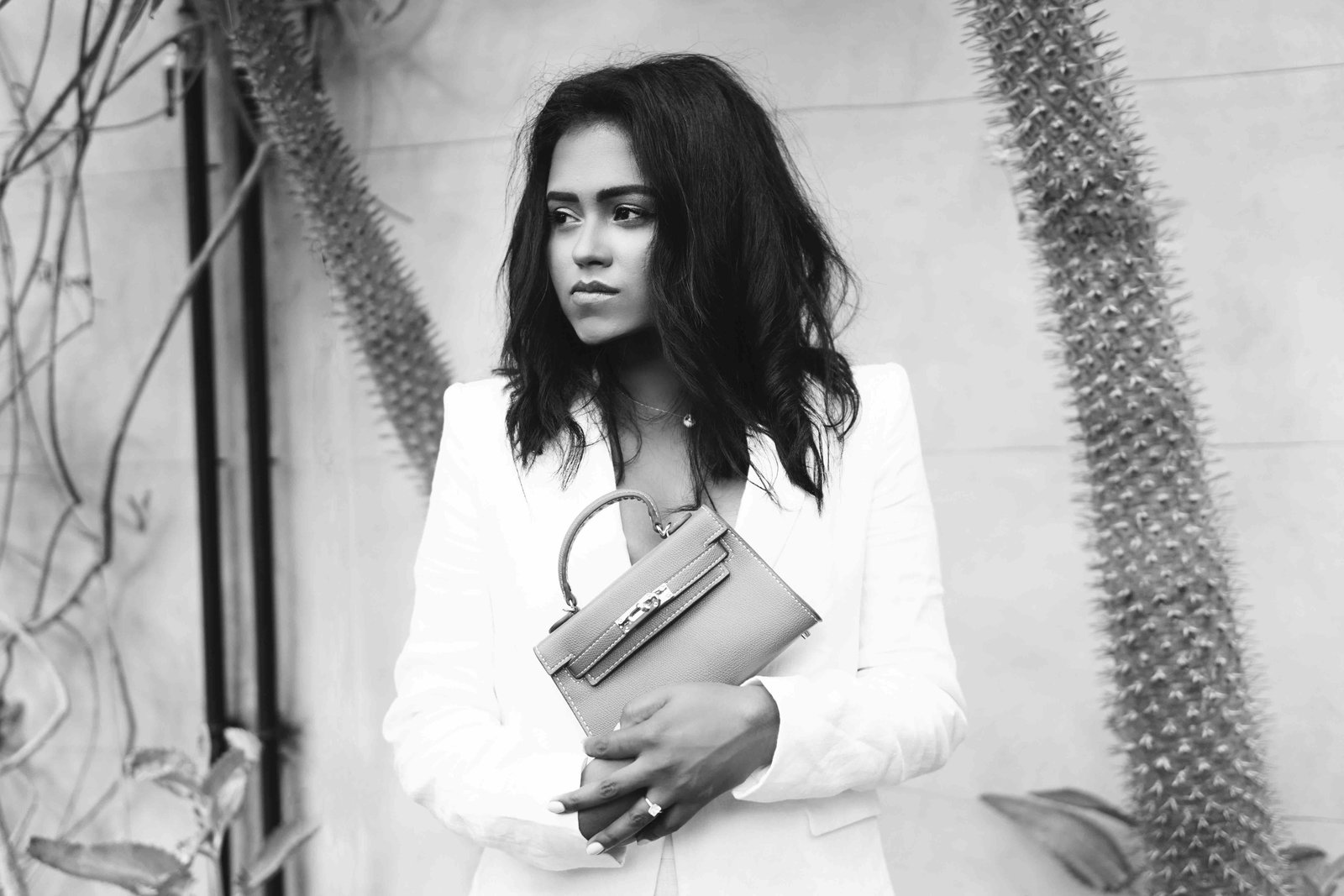 Black and white close up of Sachini wearing a white suite holding a brown Hermès mini Kelly bag