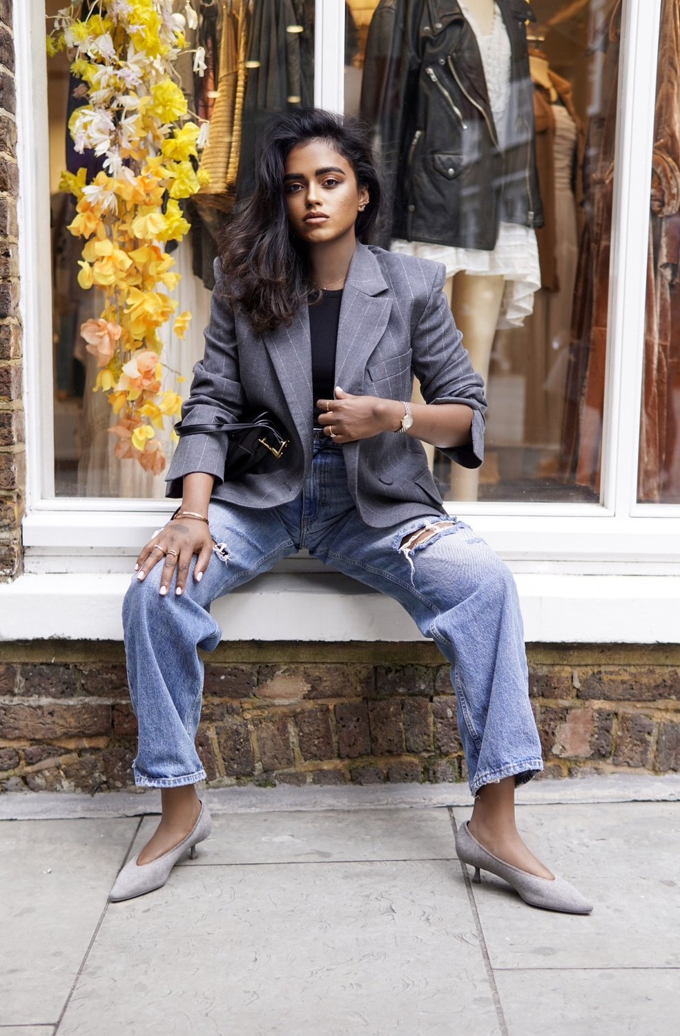 Sachini sitting in front of a shop window wearing a grey blazer, blue ripped jeans, a black Dior saddle bag and grey Vivaia shoes