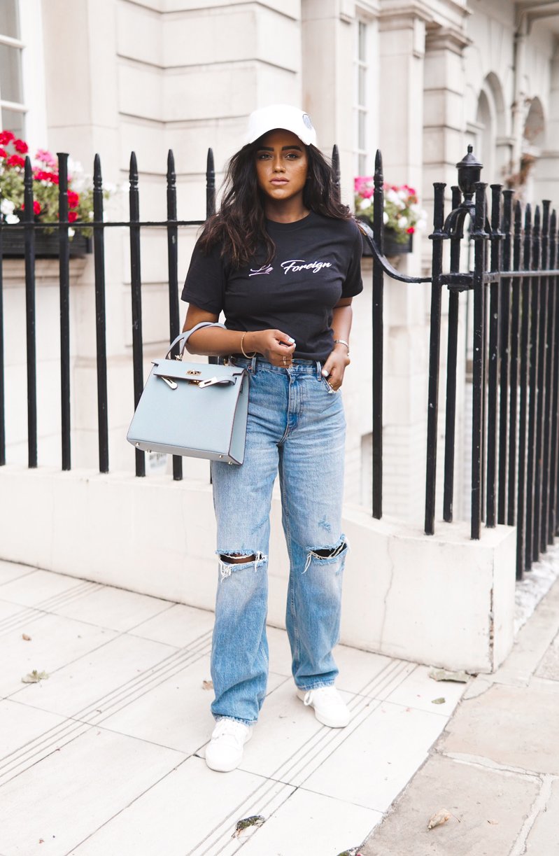 Sachini standing in front of a fence wearing a white cap, a black Le Foreign top and ripped blue jeans holding a light blue Hermès Kelly bag