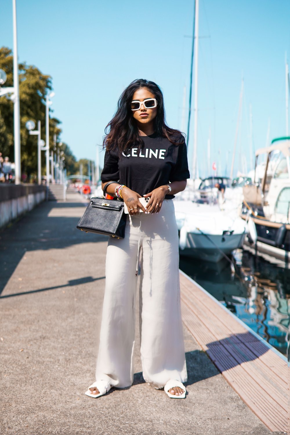 Sachini at a harbour in front of boots wearing a black Celine top and beige trousers holding a black Hermès Kelly bag