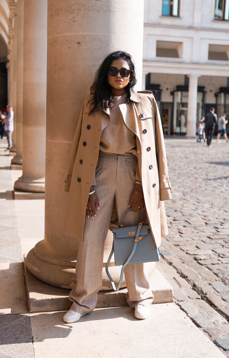 Sachini standing in front of a pillar wearing a cream colour sweater and trousers holding a blue Hermès Kelly bag