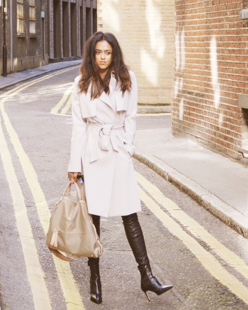 Sachini wearing a beige trench coat and black boots holding a green Prada bag