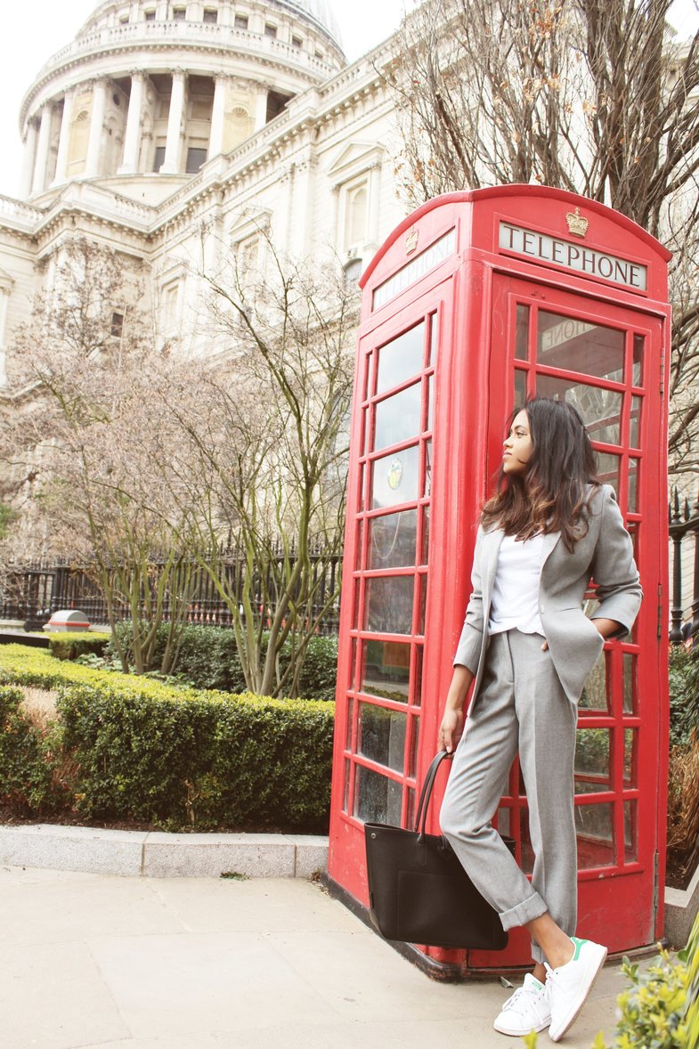 Sachini wearing a grey suit and white Adidas sneakers holding a black bag in front of a red phone booth next to St Paul