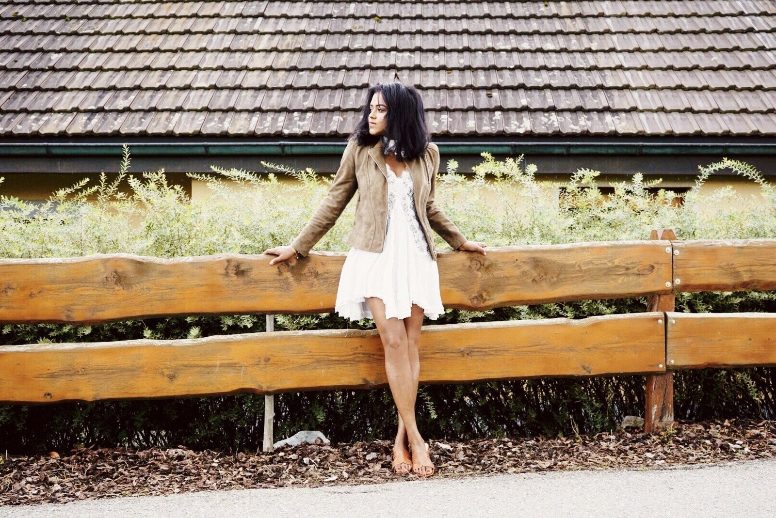 Sachini wearing a brown jacket and white dress in an outdoor environment leaning against a fence