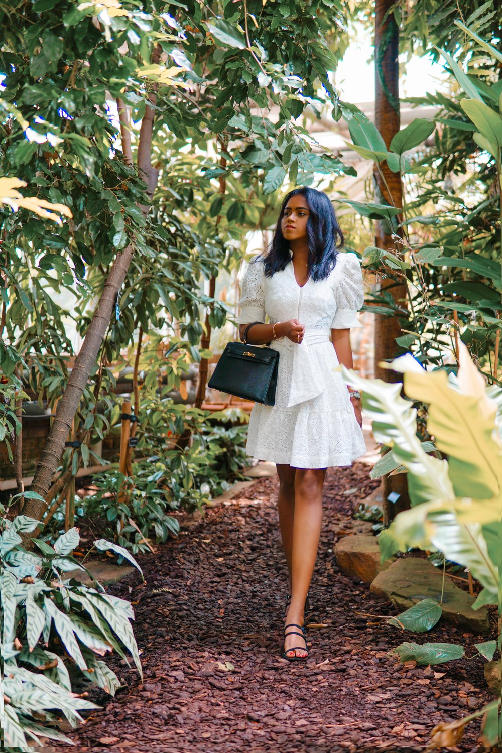 Sachini wearing a white dress holding a black Hermès Kelly bag walking in a botanic garden
