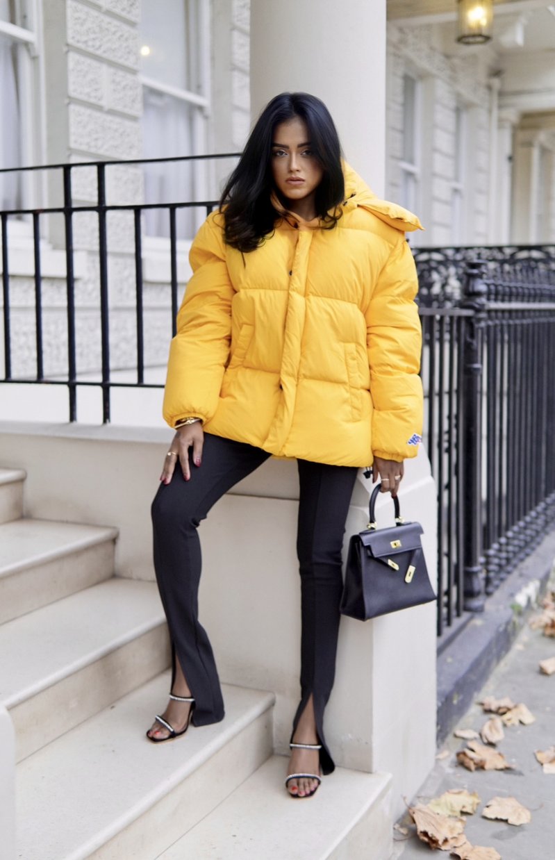 Sachini wearing a yellow Diesel puffer jacket and black Amina Muaddi shoes holding a black Hermès Kelly bag