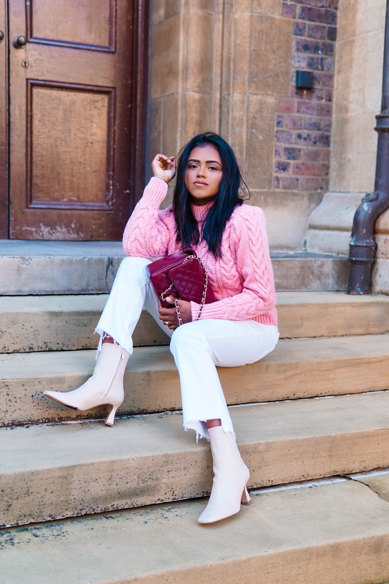 Sachini sitting on stairs wearing pink knitwear, white trousers and white boots holding a red handbag