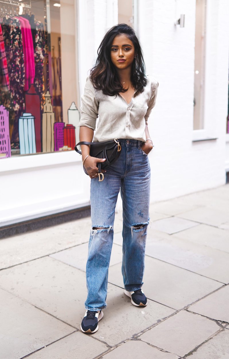 Sachini standing in front of a clothing store wearing a beige With Nothing Underneath top and blue ripped jeans holding a black Dior saddle bag