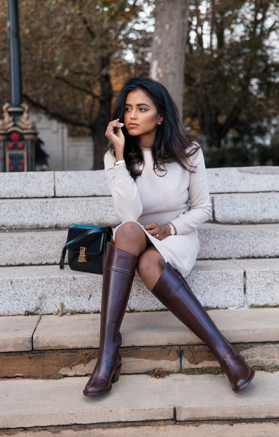 Sachini sitting on stairs wearing a beige dress and brown Fairfax and Favor high boots, with a black Celine back next to her