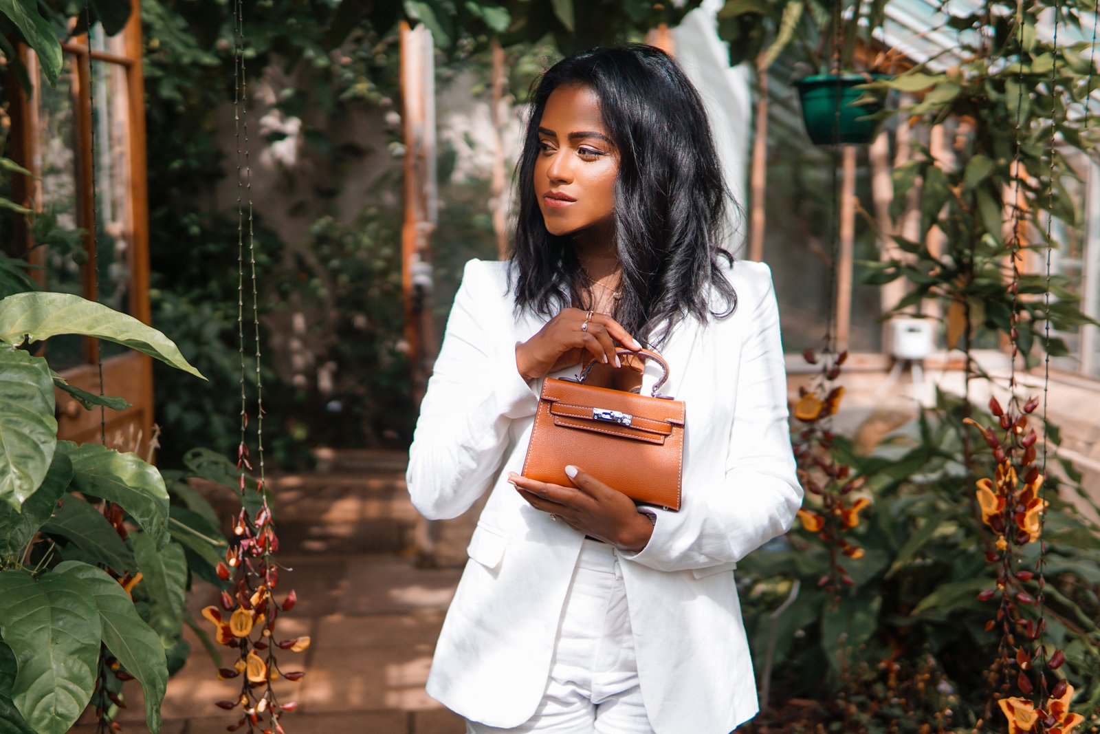 Closeup of Sachini standing wearing a white suit holding a brown Hermès mini Kelly bag