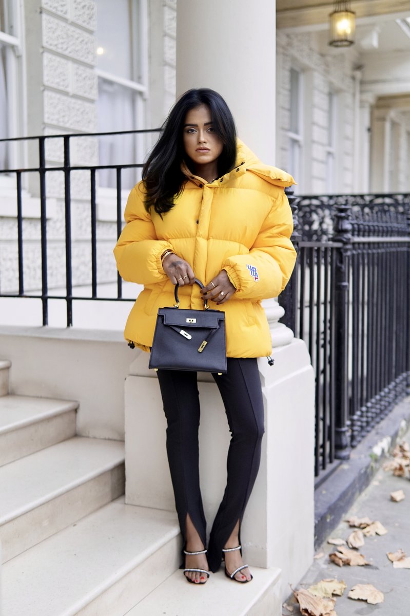 Sachini wearing a yellow Diesel puffer jacket and black Amina Muaddi shoes holding a black Hermès Kelly bag