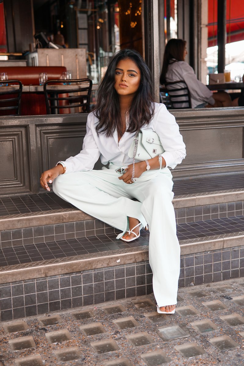 Sachini sitting on stairs in front of a cafe wearing white Bottega shoes, white top and bottom and a white Ecosusi bag