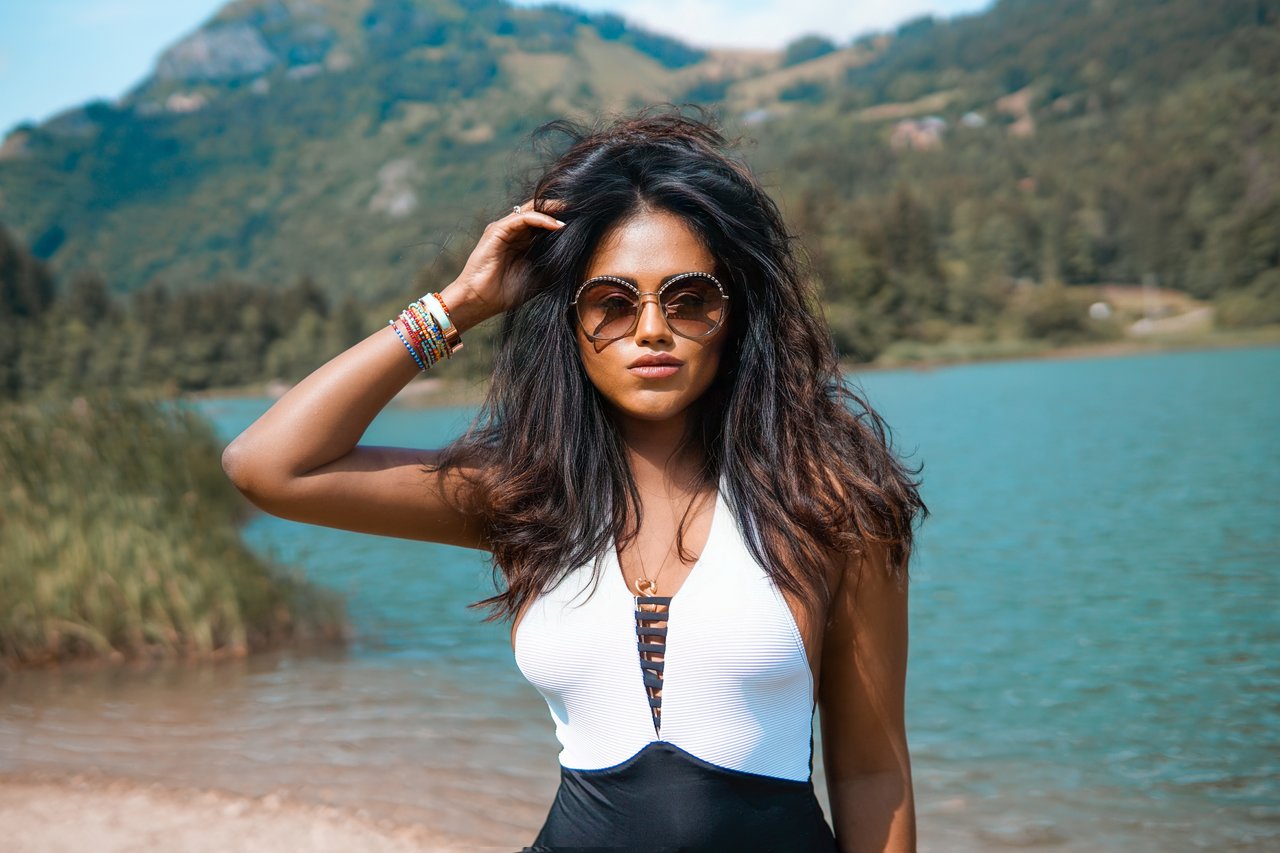 Close up of Sachini wearing Chanel sunglasses and a black and white swimsuit standing in front a mountain lake