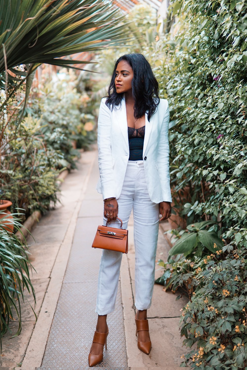 Sachini standing between exotic trees wearing a white suit holding a brown Hermès mini Kelly bag