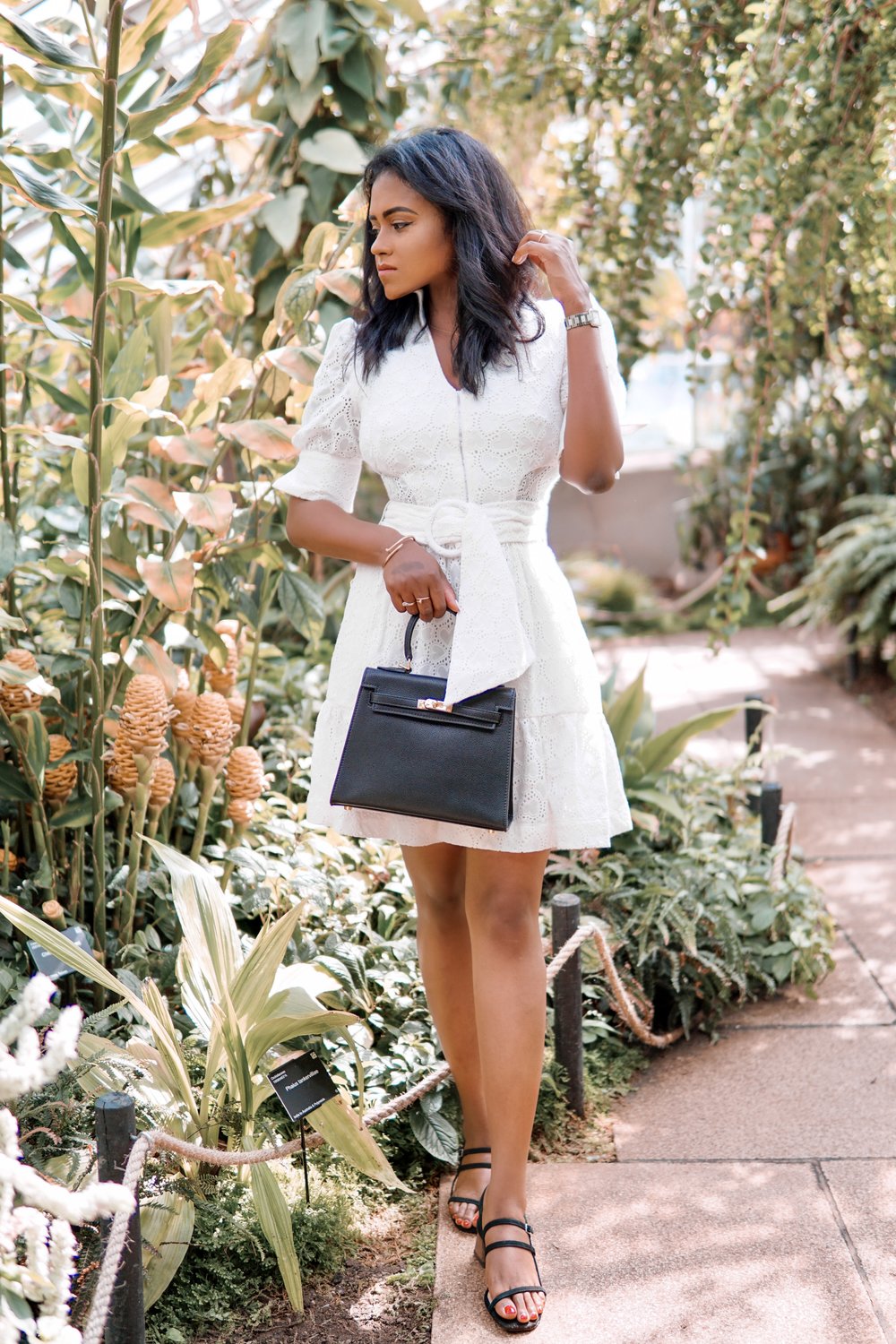 Sachini wearing a white dress holding a black Hermès Kelly bag walking in a botanic garden