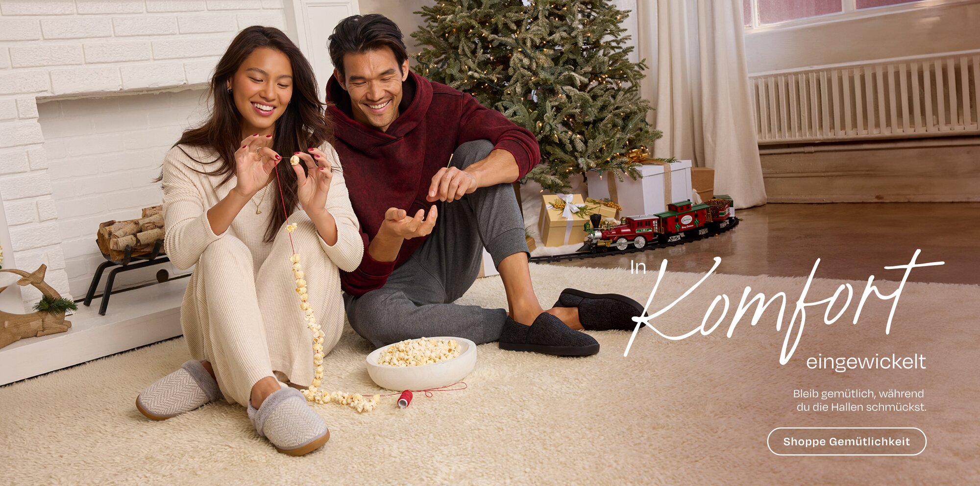 In Komfort eingewickelt Bleib gemütlich, während du die Hallen schmückst. Shoppe Gemütlichkeit. Models in Hausschuhen, die auf einem Teppich Popcorn fädeln.