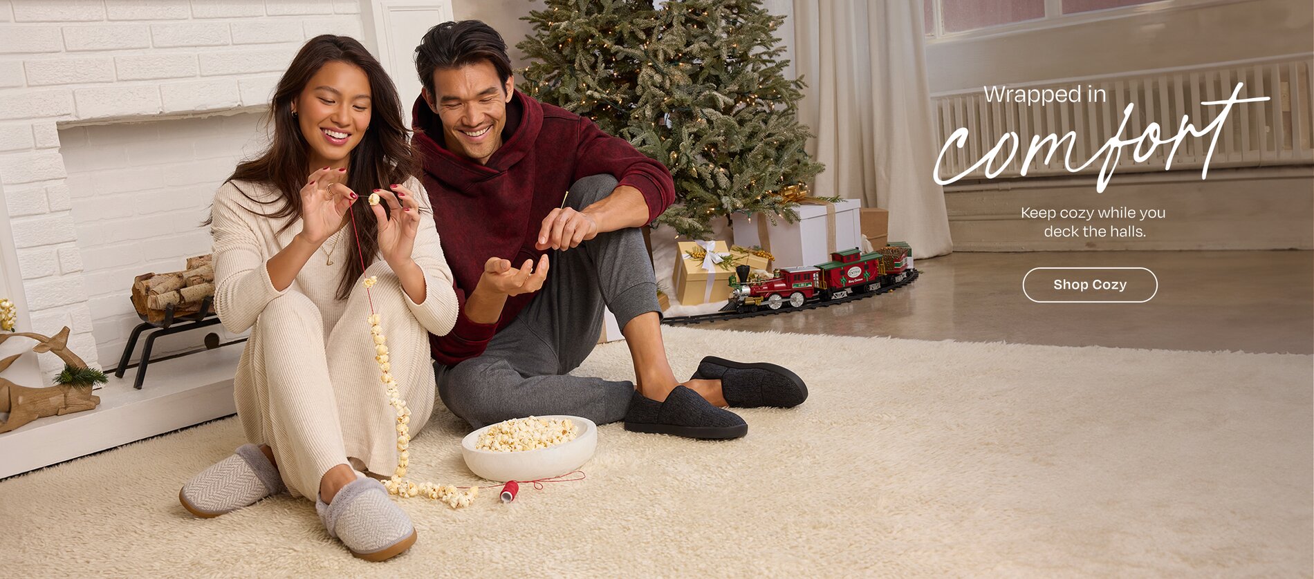 Wrapped in comfort. Keep cozy while you 
deck the halls. Shop Cozy. Models in slippers sitting on rug stringing popcorn.