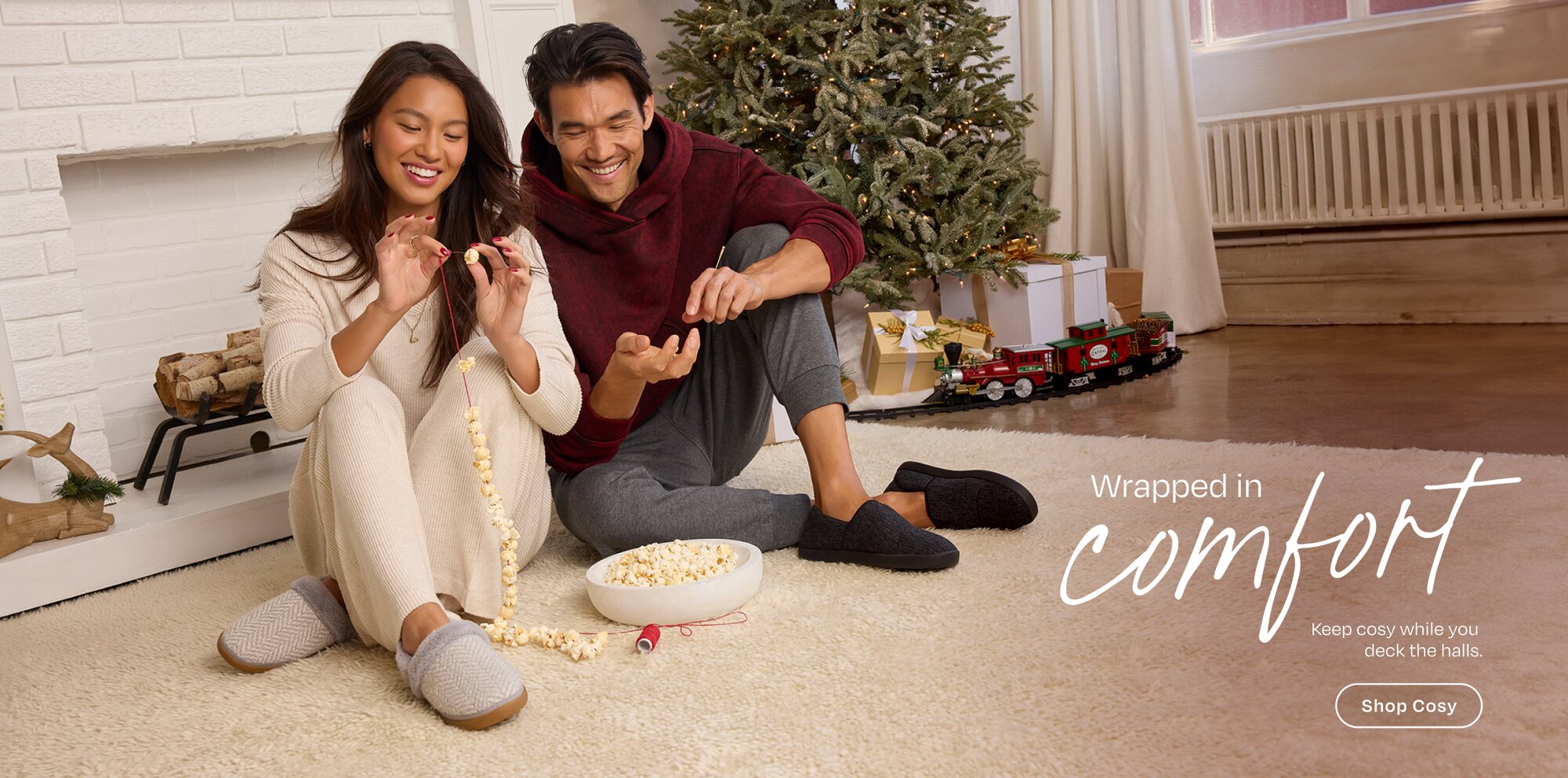 Wrapped in comfort. Keep cosy while you deck the halls. Shop Cosy Models in slippers sitting on rug stringing popcorn.