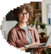 Smiling woman with curly hair and glasses, wearing a brown shirt, using a laptop in a home office setting