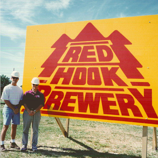 Two men in hard hats stand in front of yellow and red Redhook Brewery sign