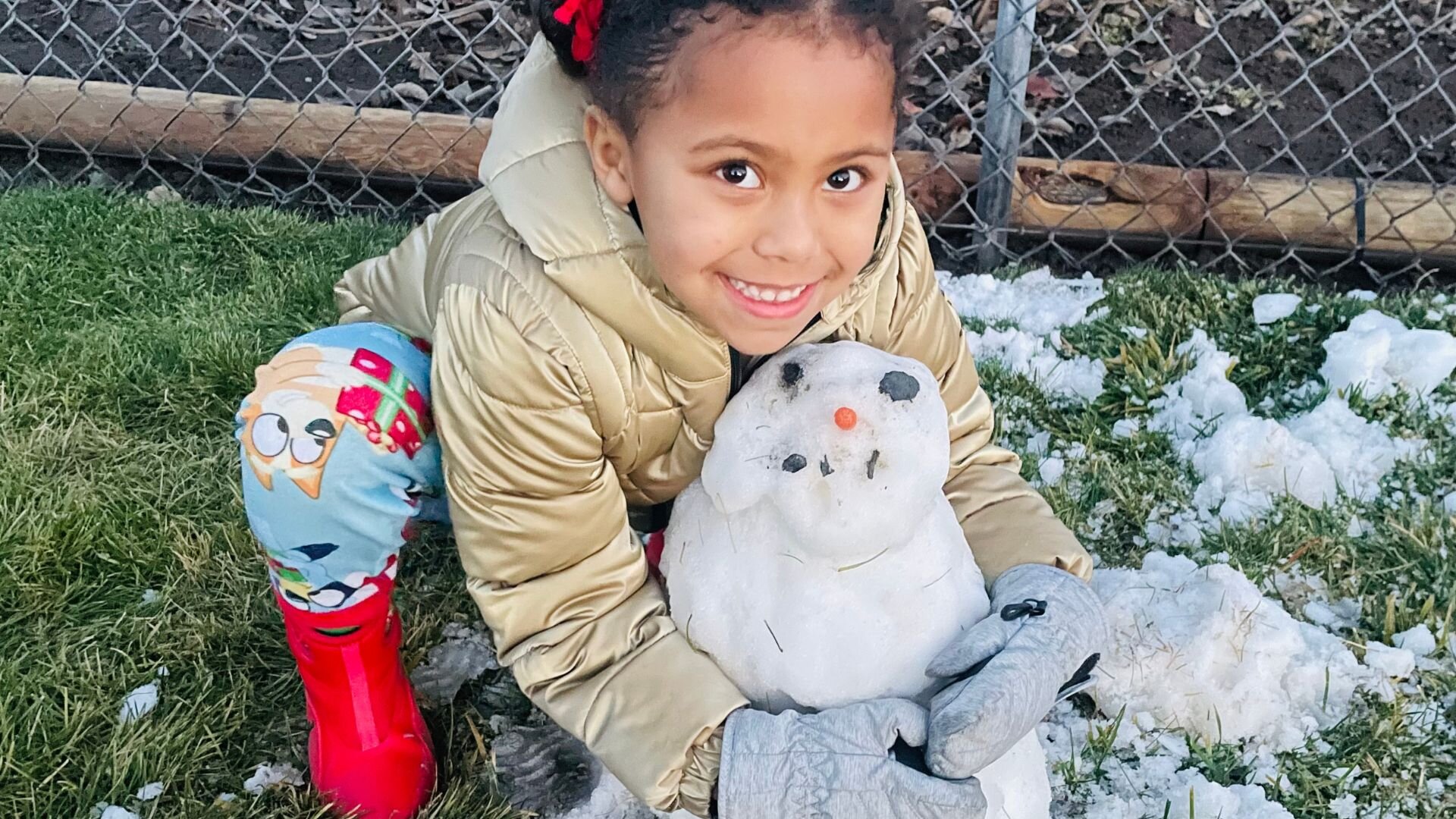 A little girl hugging the small snowman she made in her yard.
