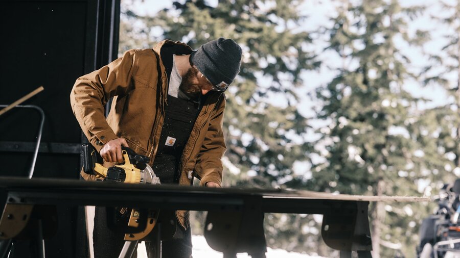 A man cutting wood in his barn wearing a winter work coat and beanie.