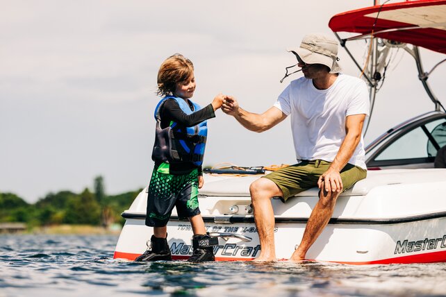 A young wakeboarder being encouraged by an older coach on the back of a wakeboard boat