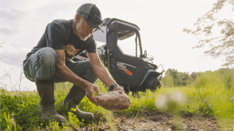 A man near a side-by-side bends down to drop a Redmond Trophy Rock on a deer mineral site.