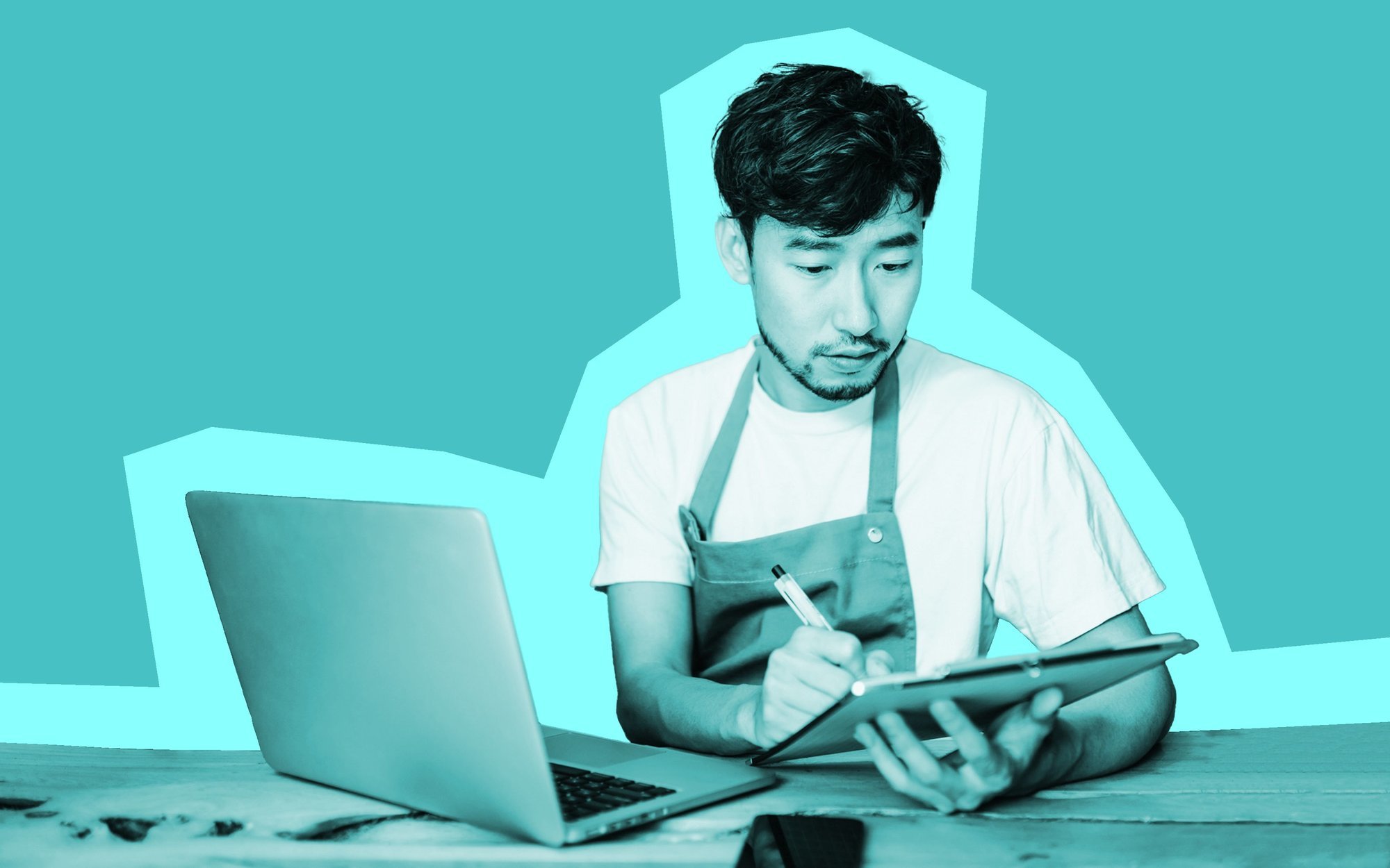 Black and white photo of a person wearing an apron and holding a tablet seated at a work bench in front of a laptop.