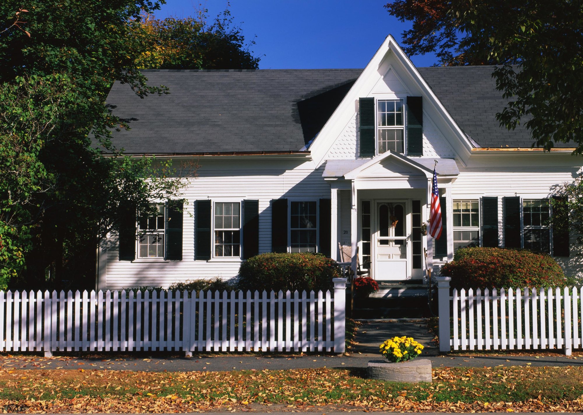 White picket fence in front of white colonial home with American flag